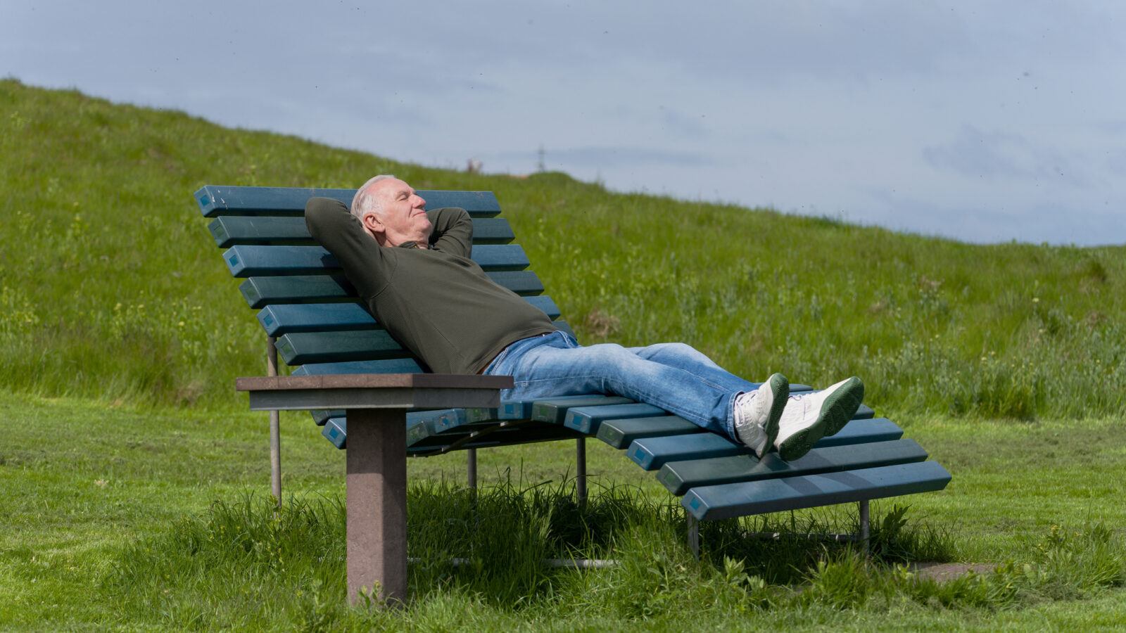 Älterer Mann, der auf einer Bank im Sonnenlicht auf einer Wiese liegt.